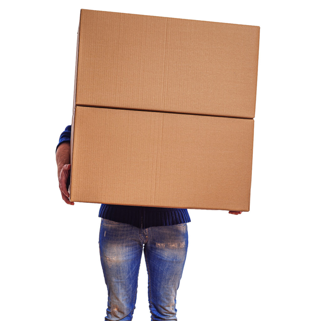 a person wearing jeans and boots stands on a wooden floor while holding two large cardboard boxes that hide their upper body.